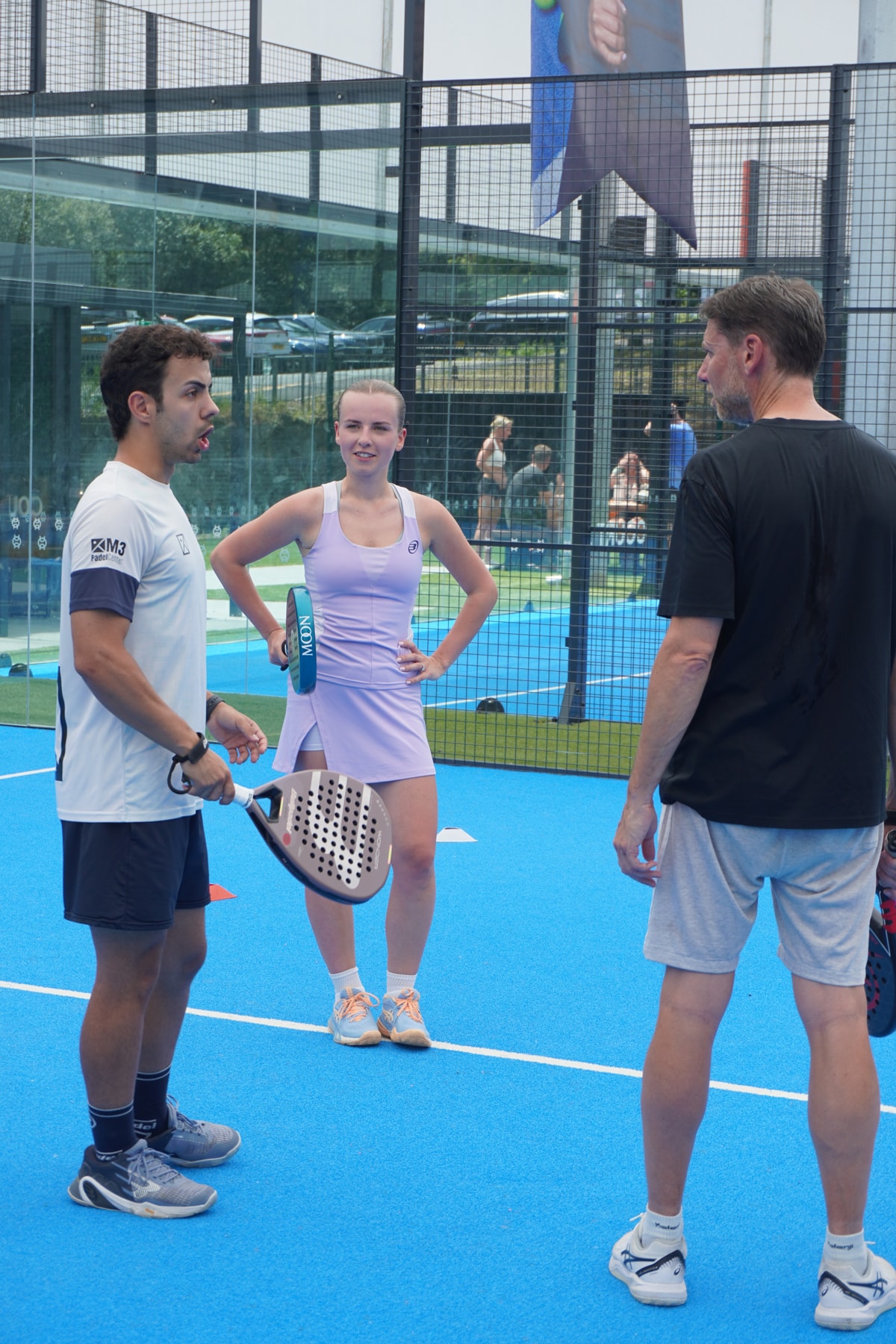 A coach in a white M3 shirt talks two players through a drill on a blue padel court with glass walls behind